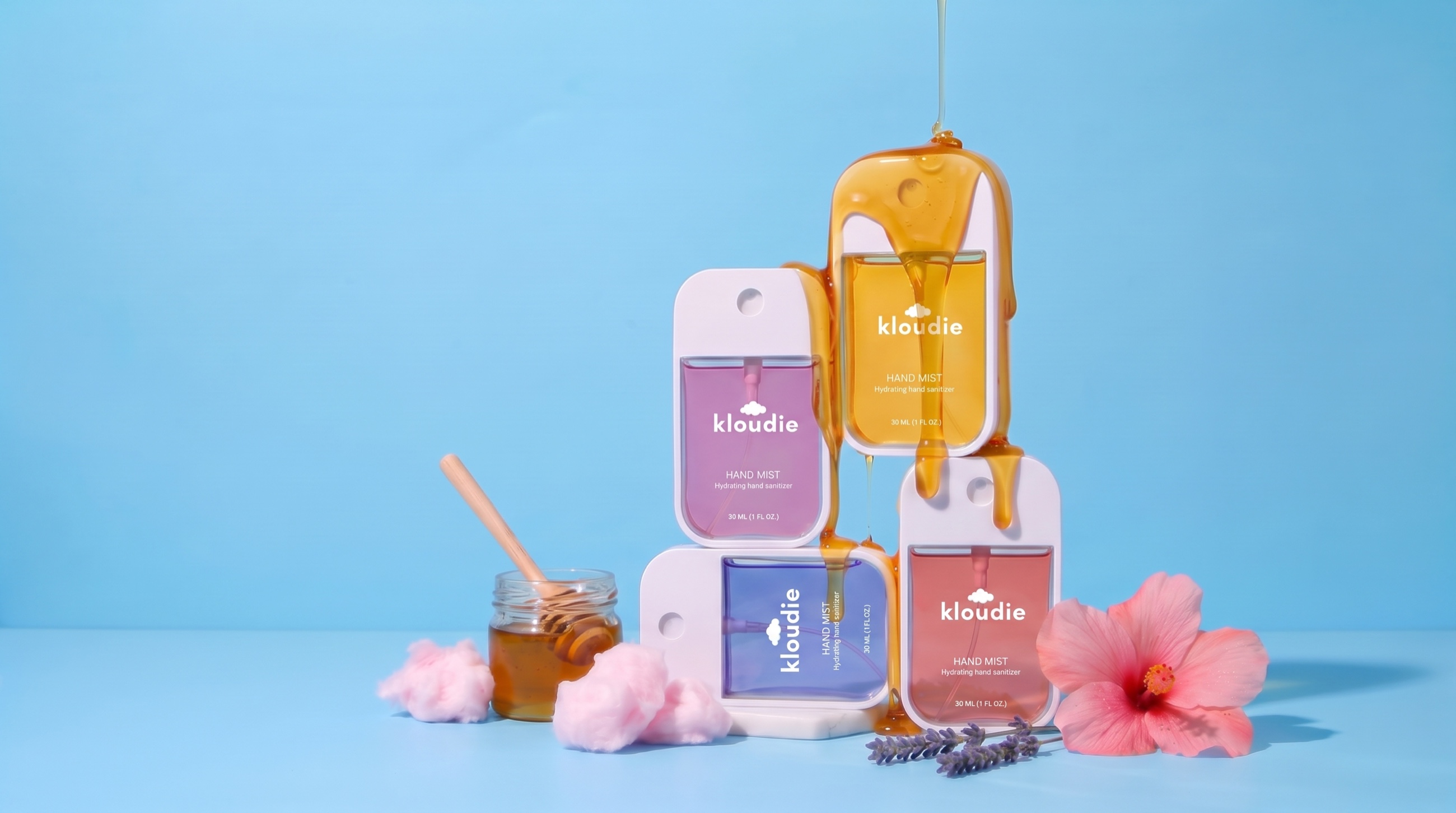 Kloudie hand mist lineup in four scents on blue, with honey pouring over the bottles and cotton candy, lavender and hibiscus styled around them